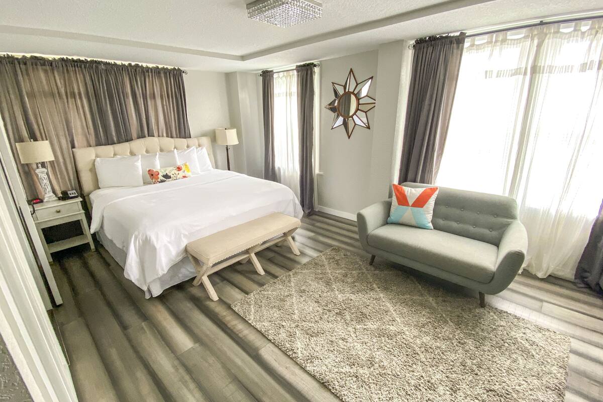 A hotel room with a large bed, a bedside table with a lamp, a sofa with a cushion, and a decorative mirror on the wall.