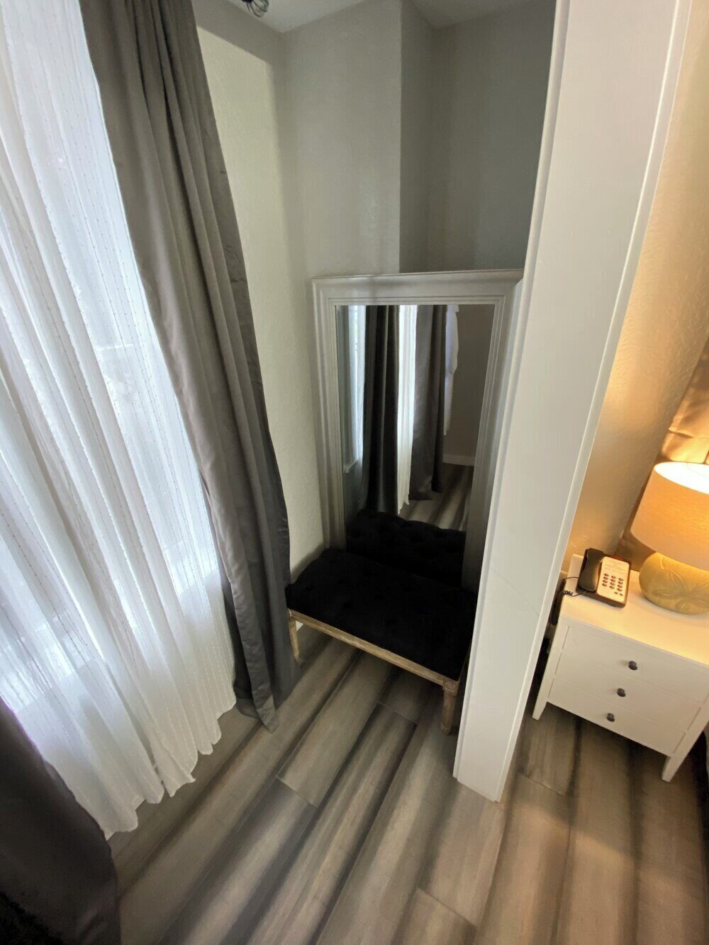 A narrow room with a bed, a nightstand with a lamp, and a door leading to another room.