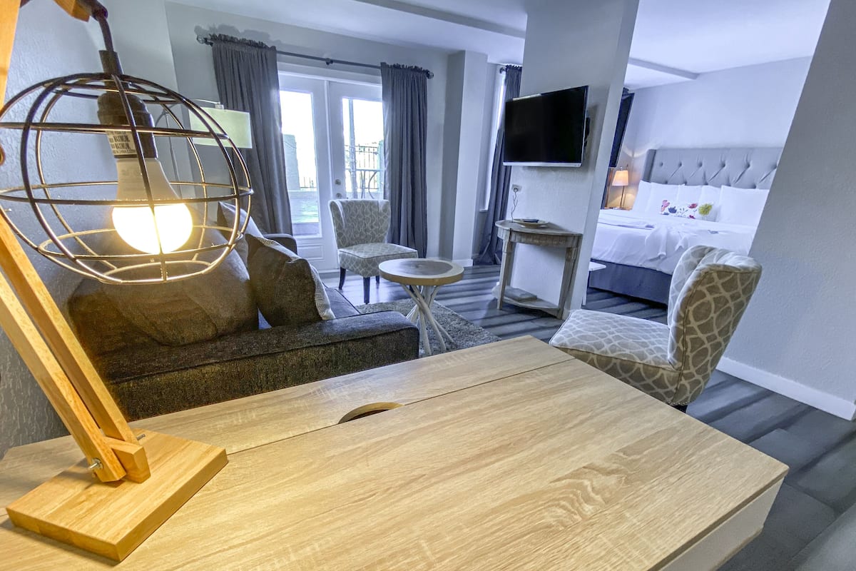 A hotel room with a wooden desk, a sofa, a patterned armchair, a small round table, a television mounted on the wall, and a bed with a grey headboard.