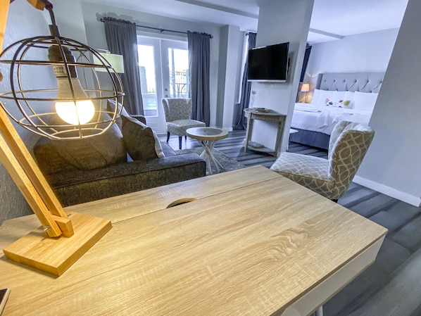 A hotel room with a wooden desk, a sofa, a patterned armchair, a small round table, a television mounted on the wall, and a bed with a grey headboard.
