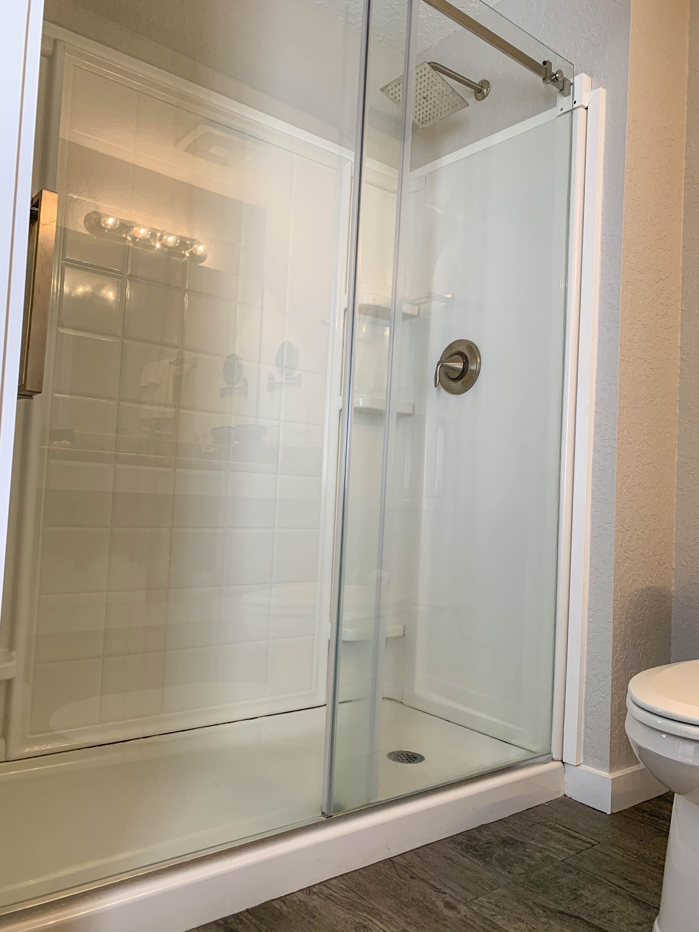 A glass-enclosed shower with a white tiled wall and a white toilet in a bathroom.