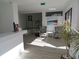A modern reception area with a wooden floor, a reception desk, two patterned armchairs, a potted plant, and a wall-mounted shelf with bottles.