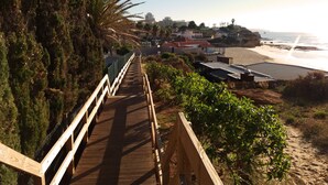 Eine Holzpromenade führt zu einem Strand, im Hintergrund sind Gebäude zu sehen.