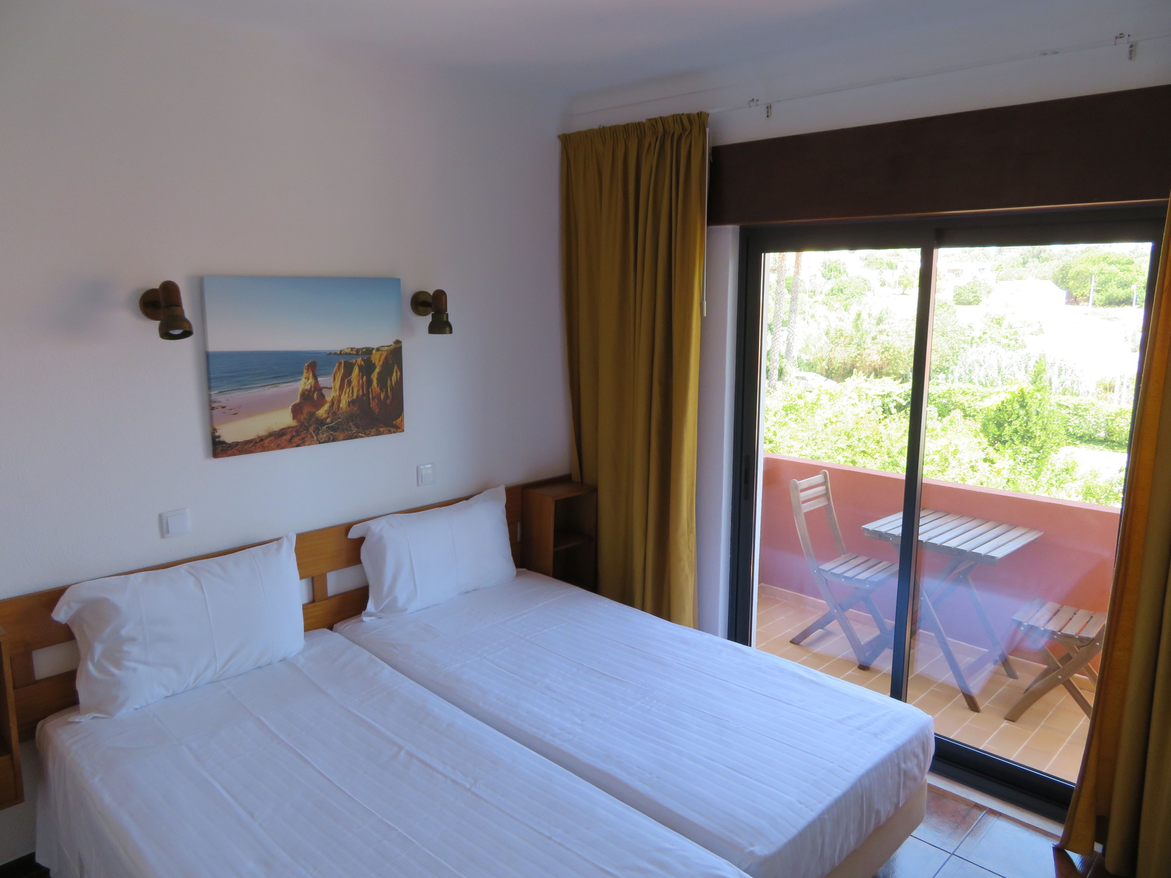 Uma cama de casal com roupa de cama branca, um quadro de uma paisagem de praia, uma porta de correr de vidro para uma área externa com uma mesa e cadeiras, e uma cortina marrom.