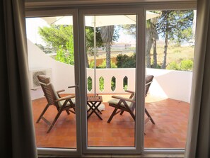 Eine Terrasse mit zwei Holzstühlen und einem Tisch, einer weißen Wand und Blick auf Grünflächen und Gebäude.