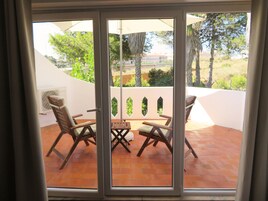 Un patio con dos sillas de madera y una mesa, una pared blanca y vistas a zonas verdes y edificios.