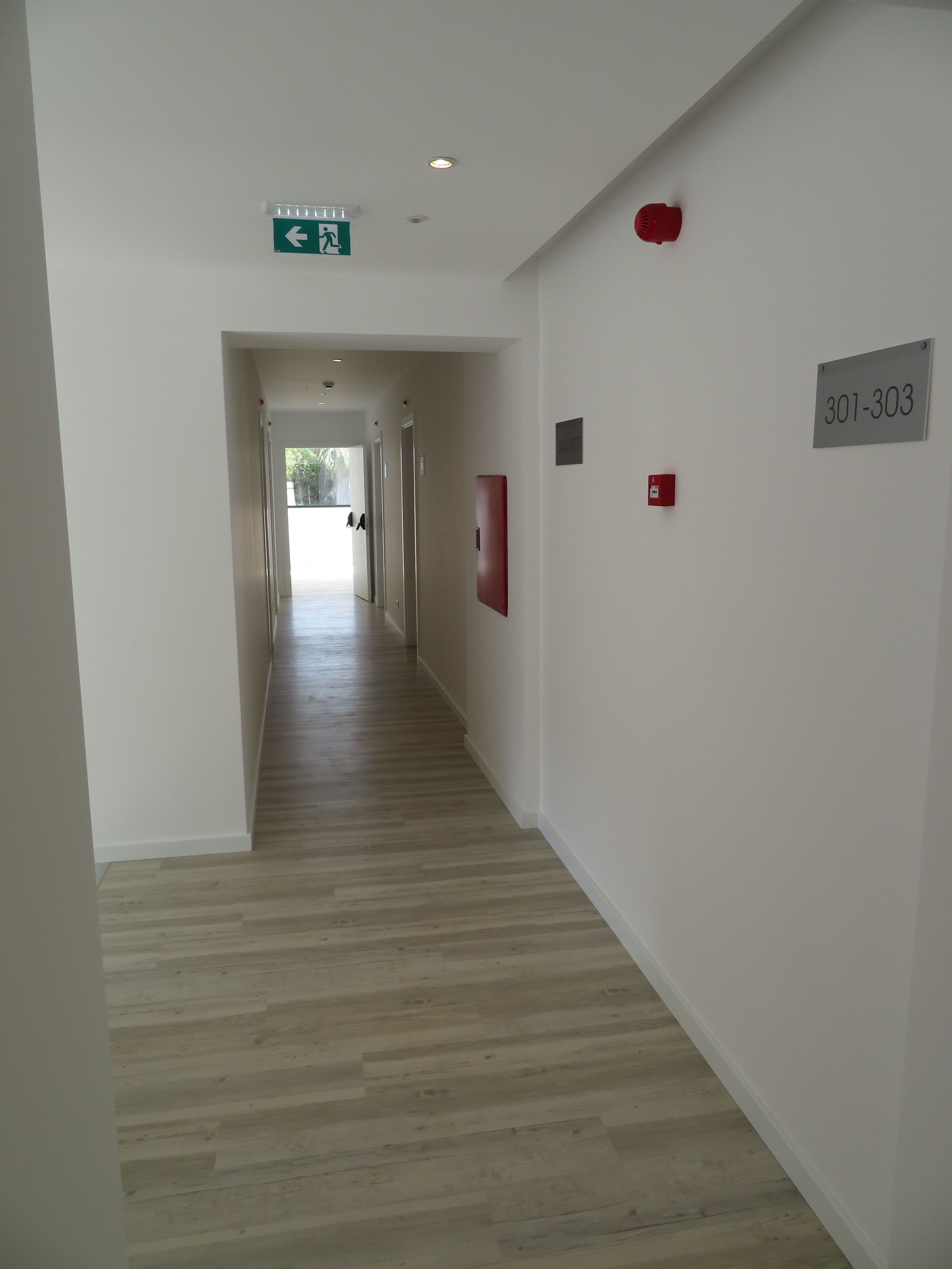 Um corredor com piso de madeira, uma placa de saída de emergência verde, um alarme de incêndio vermelho e uma placa com os números 301-303.