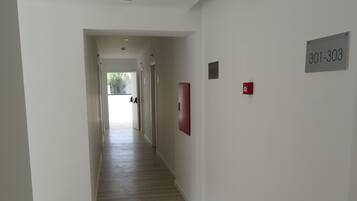 Um corredor com piso de madeira, uma placa de saída de emergência verde, um alarme de incêndio vermelho e uma placa com os números 301-303.