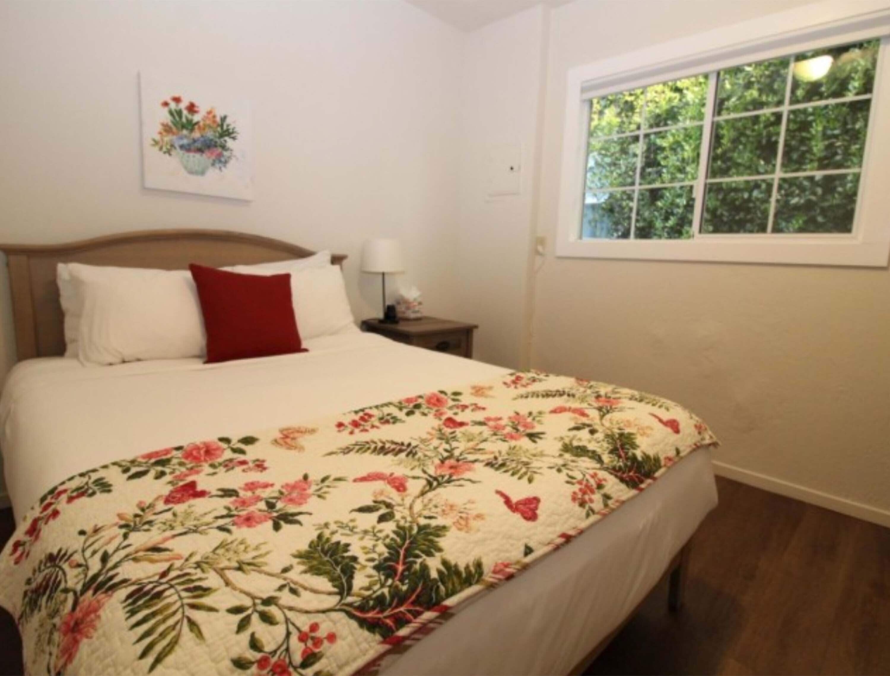 Une chambre à coucher avec une tête de lit en bois, une couvre-lit à motifs floraux, un oreiller rouge, une table de chevet avec une lampe et une fenêtre donnant sur un paysage verdoyant.