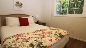 Une chambre à coucher avec une tête de lit en bois, une couvre-lit à motifs floraux, un oreiller rouge, une table de chevet avec une lampe et une fenêtre donnant sur un paysage verdoyant.