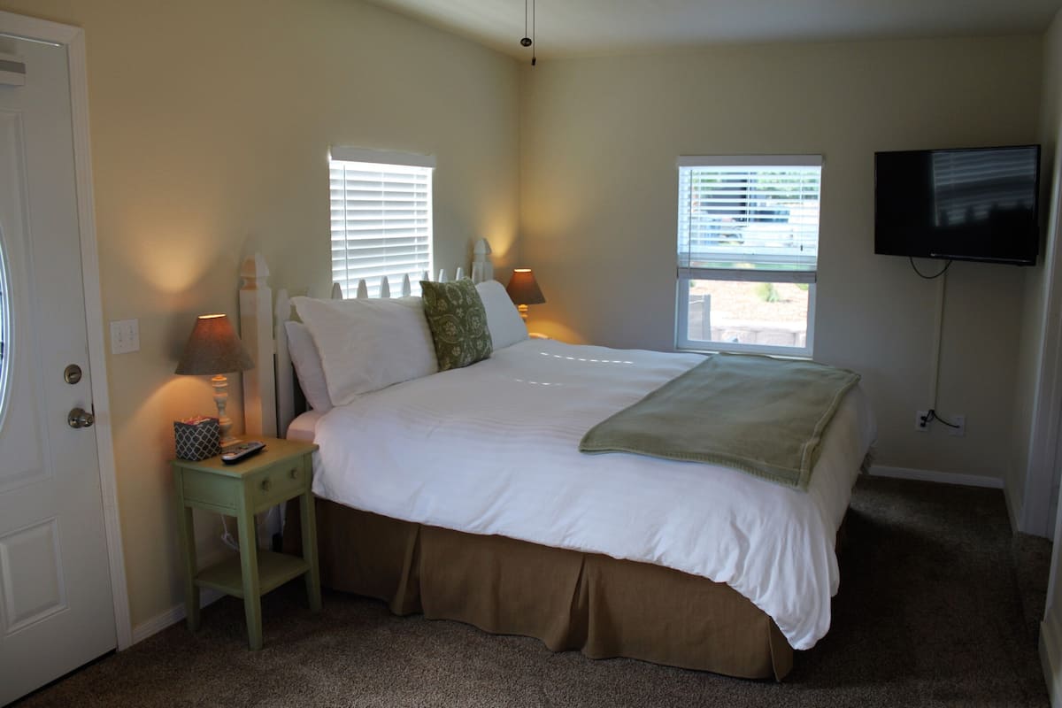 A bedroom with a bed, two bedside lamps, a TV mounted on the wall, and a window with blinds.