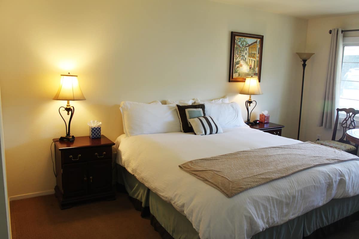 A neatly made bed with white linens, two bedside tables with lamps, a painting on the wall, and a window with curtains.