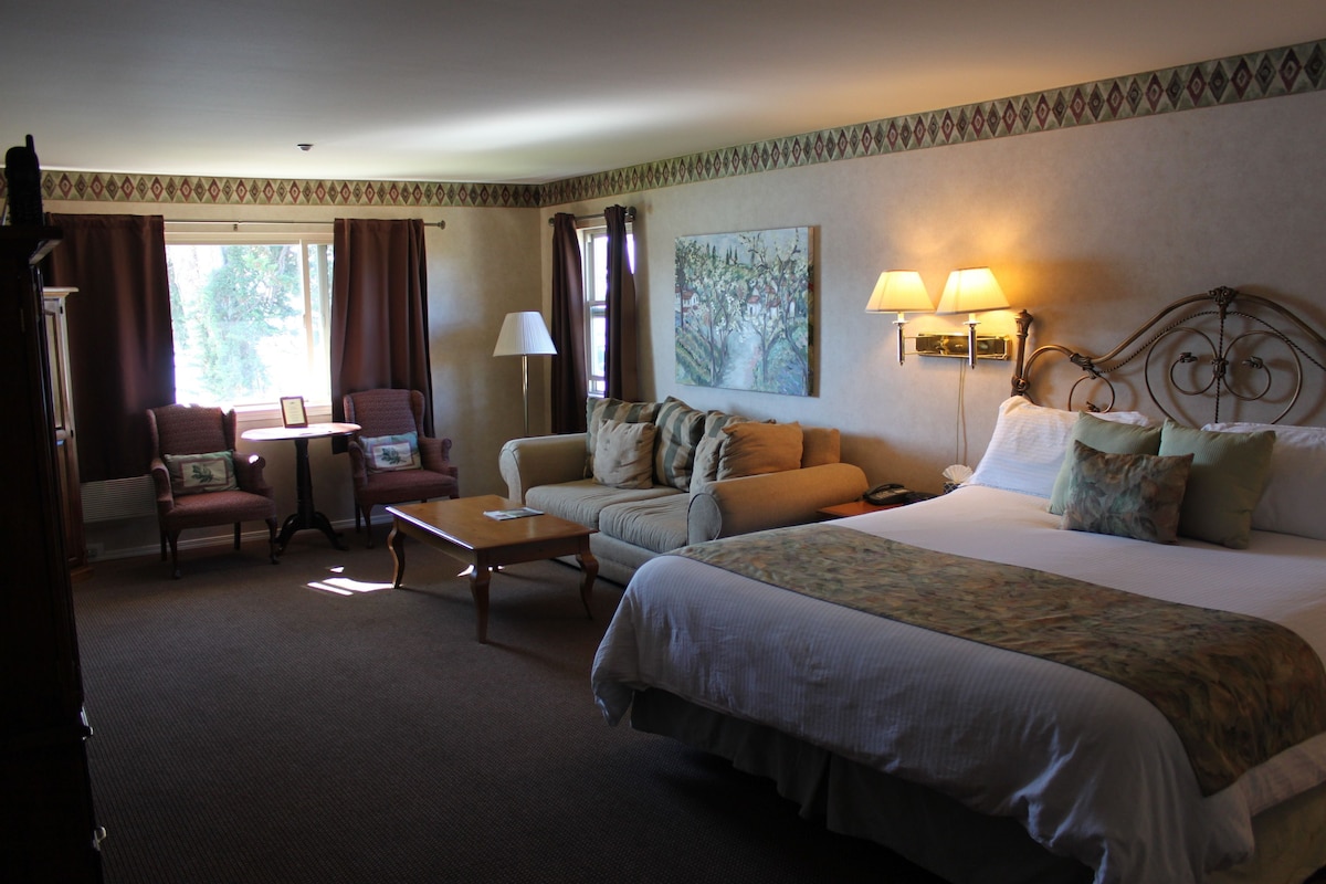 A hotel room with a large bed, two armchairs, a small table, a lamp, and a painting on the wall.
