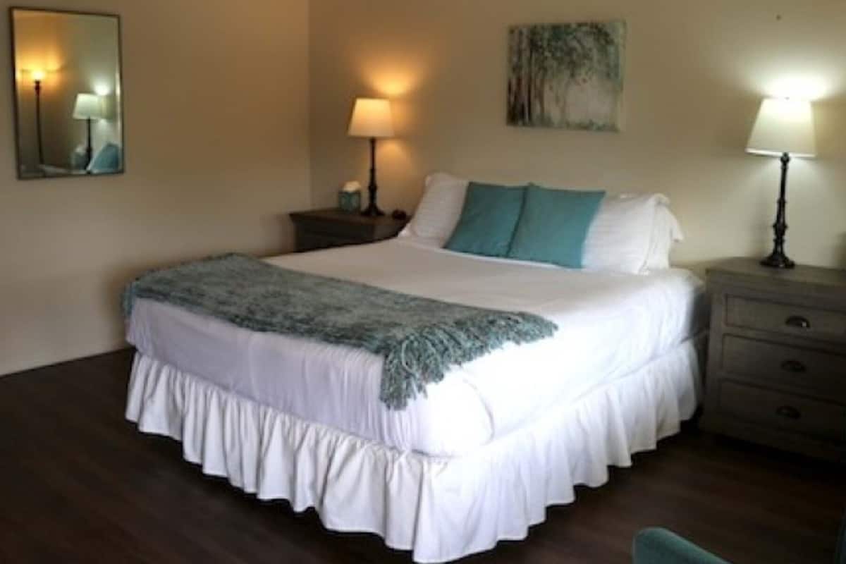 A neatly made bed with white bedding and a blue throw, a bedside table with a lamp, and a wall-mounted painting.