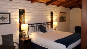 A bedroom with a wooden ceiling, white brick walls, a bed with white bedding, two bedside tables with lamps, and a chair. - Back Bay Inn (Los Osos)