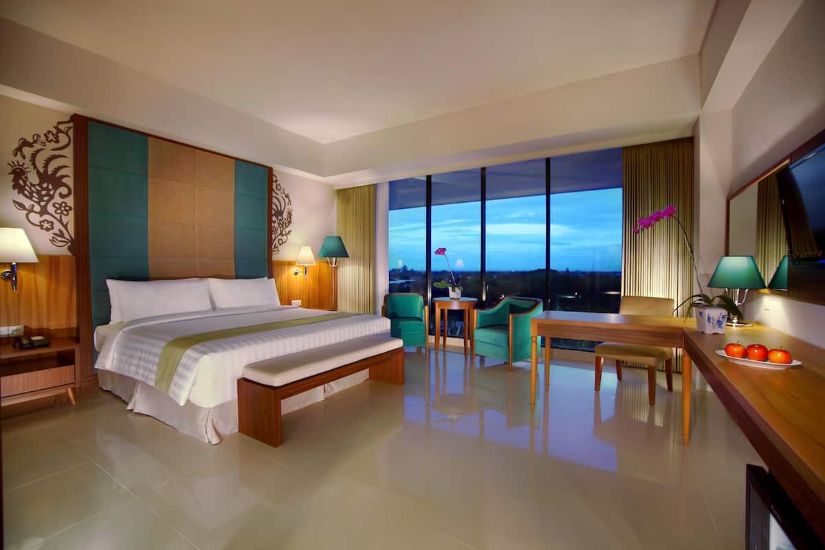 Een hotelkamer met een groot bed, twee stoelen, een kleine tafel, een bureau met een schaal fruit en een groot raam met uitzicht over de stad.