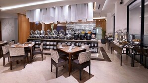 Een moderne eetruimte met meerdere tafels en stoelen, een centraal buffet met diverse gerechten, en een keuken in de achtergrond.
