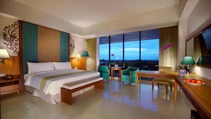 Hotelkamer met een groot bed, een bureau, twee stoelen, een bijzettafeltje en uitzicht op de stad in de schemering.