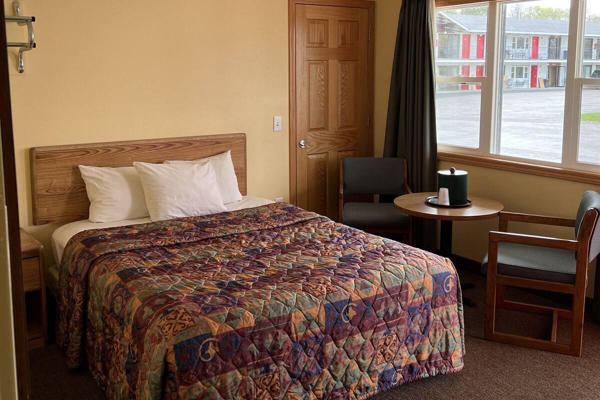 Une chambre d’hôtel avec un lit, une petite table et une chaise, une fenêtre avec des rideaux et une porte.