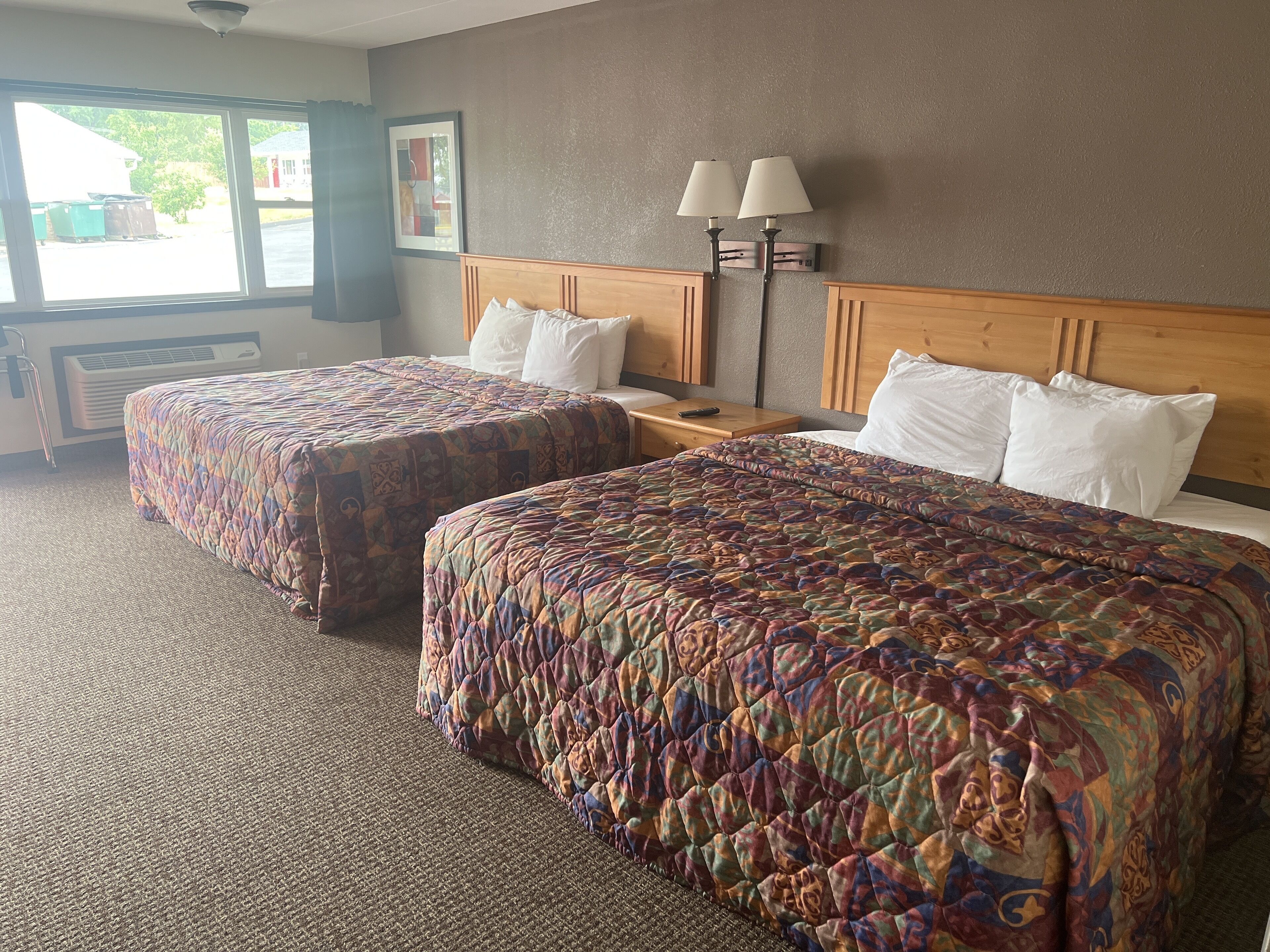 A hotel room with two queen beds, a nightstand, a lamp, and a window with a view of the outside.
