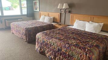 A hotel room with two queen beds, a nightstand, a lamp, and a window with a view of the outside.