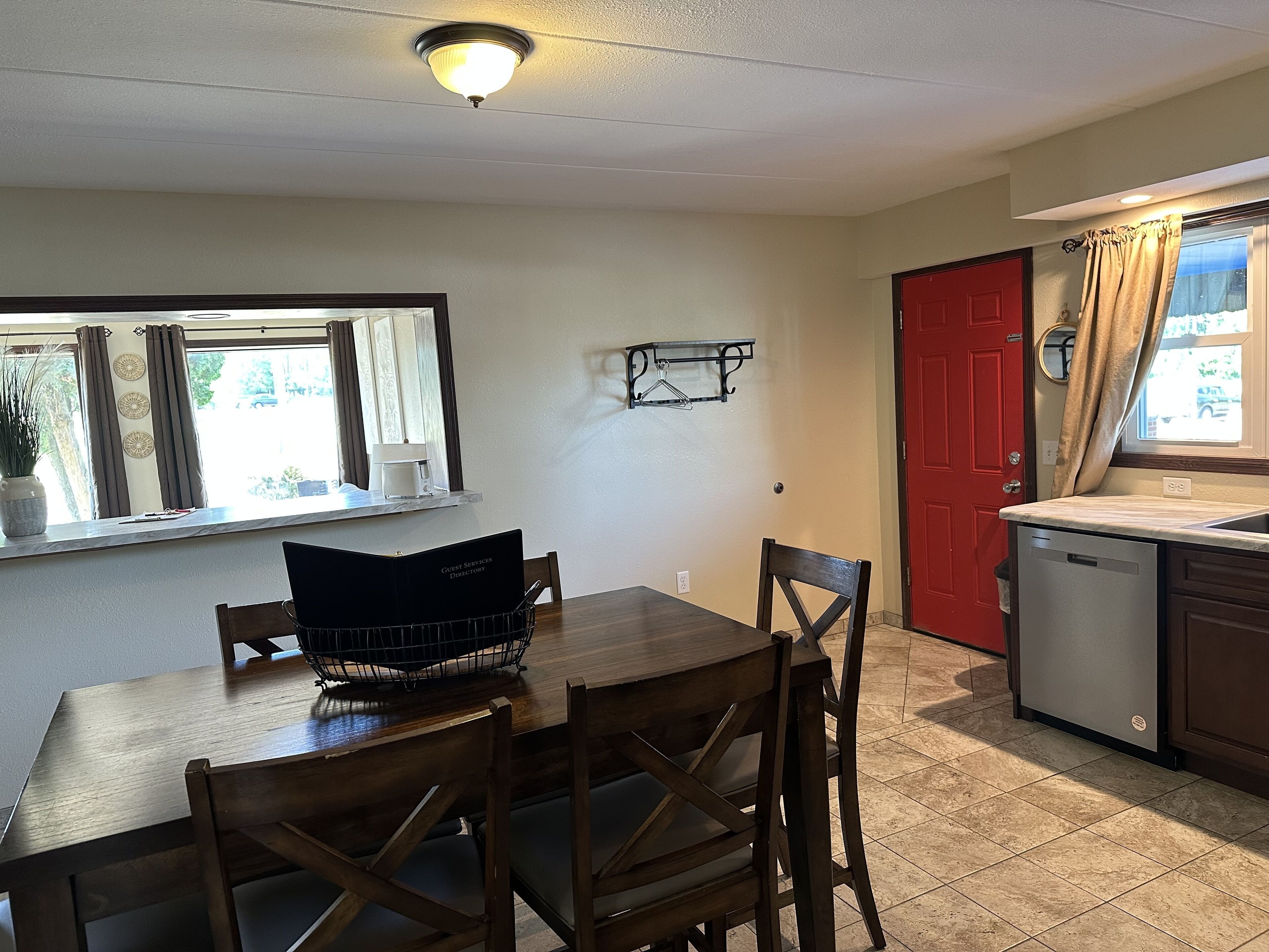 A dining area with a wooden table and chairs, a guest services directory, a window with curtains, a red door, and a kitchen area with a dishwasher.