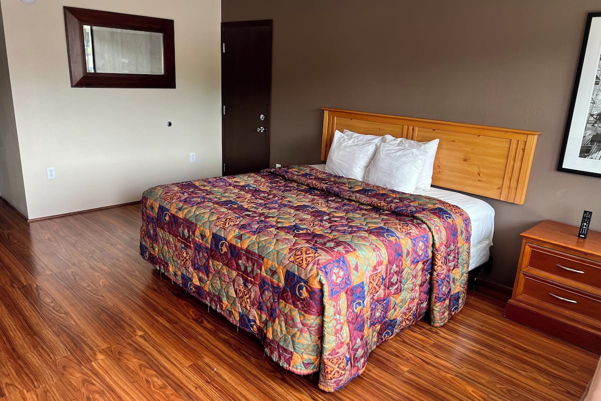 Une chambre d’hôtel avec un plancher en bois, un lit recouvert d’une courtepointe aux couleurs vives, une tête de lit en bois, une table de chevet avec une télécommande et un tableau encadré accroché au mur.