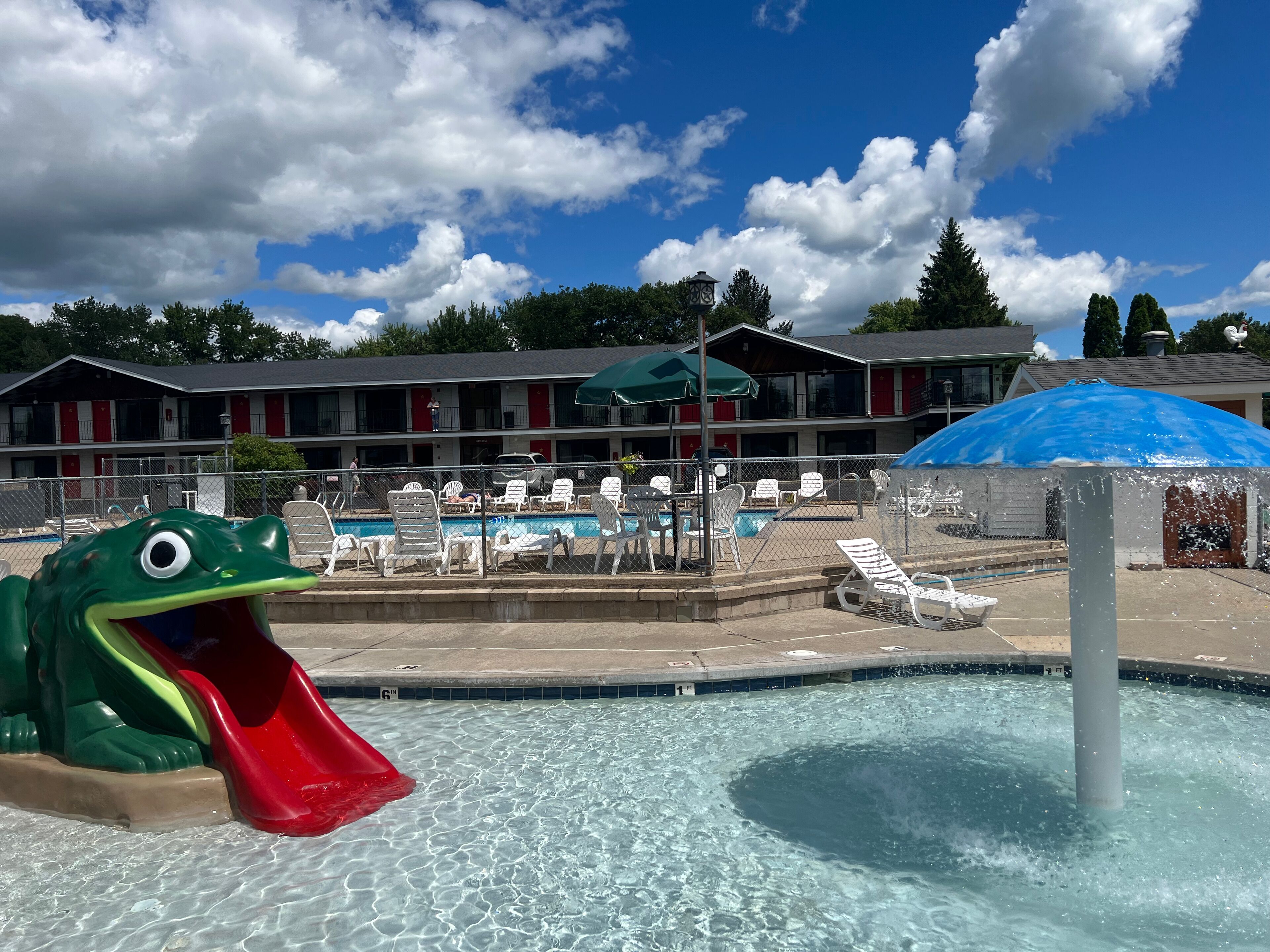 Uma área de piscina com escorregador, cadeiras de descanso e um edifício com torre do relógio ao fundo.