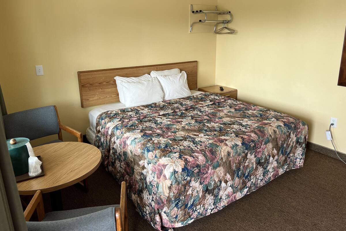Une chambre d’hôtel avec un lit, une tête de lit en bois, une table de chevet avec une télécommande, une chaise et une petite table sur laquelle se trouve une boîte de mouchoirs.