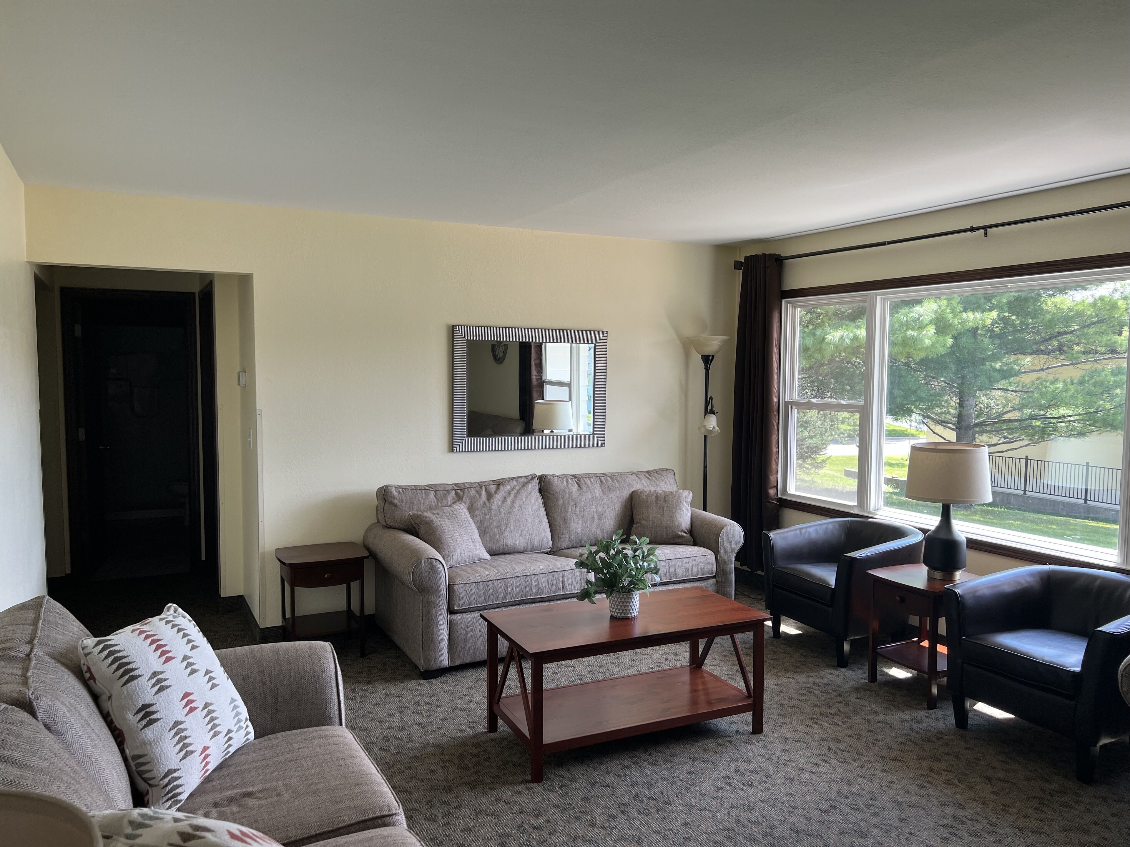 A living room with a sofa, armchair, coffee table, and a lamp. There is a window with a view of trees and a lamp on a side table.