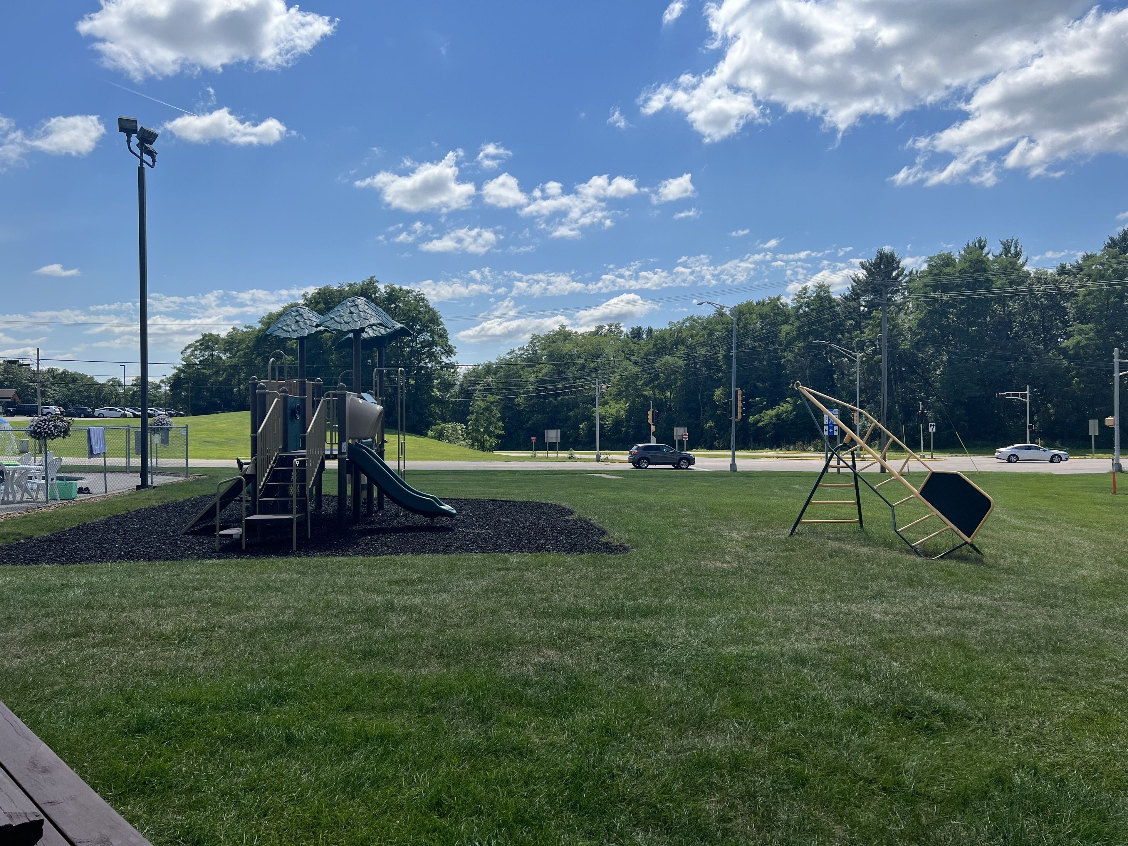 A playground with a slide and climbing structures, surrounded by a grassy area and trees.