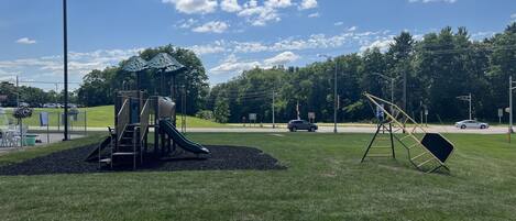 A playground with a slide and climbing structures, surrounded by a grassy area and trees.