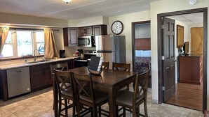A kitchen with a dining table, chairs, a microwave, a refrigerator, and a clock on the wall.