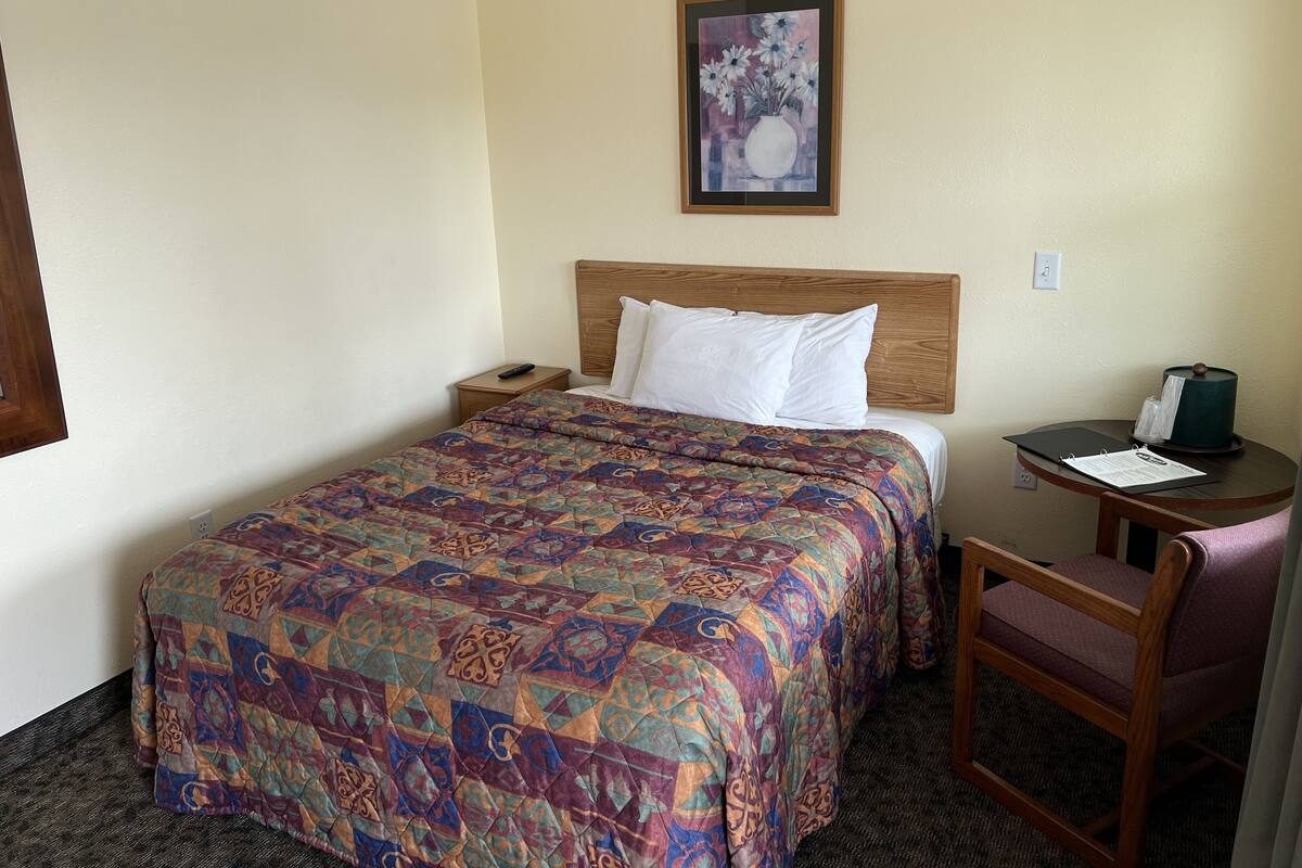 Une chambre d’hôtel avec un grand lit, une tête de lit en bois, une petite table de chevet avec une télécommande, une chaise et un bureau avec une lampe et un carnet.