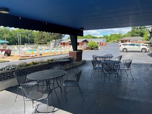 An outdoor patio with metal tables and chairs, a brick wall, and a view of a parking lot and buildings in the background.