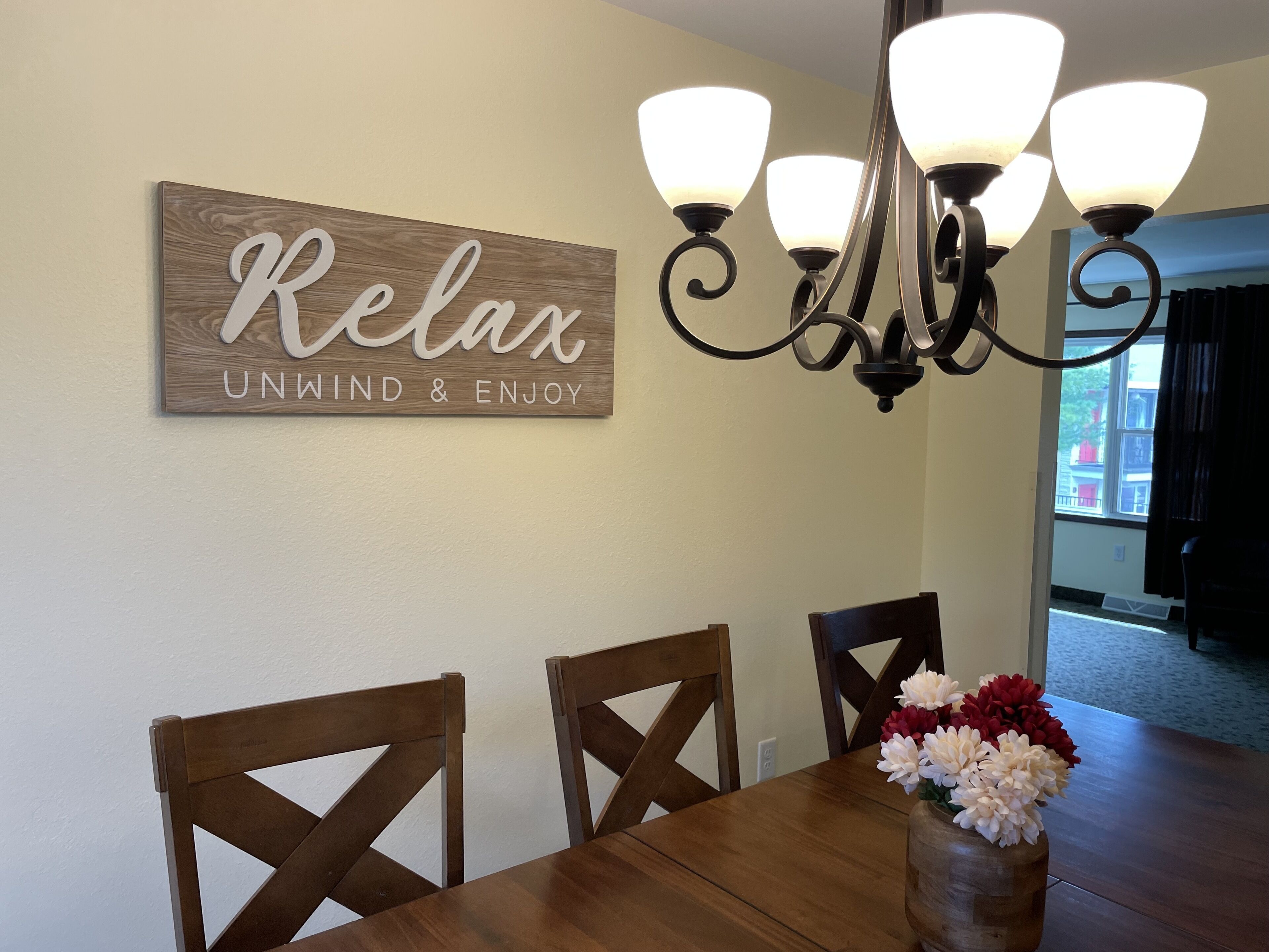 Un comedor con una mesa de madera, cuatro sillas, una araña y un letrero que dice "Relax Unwind & Enjoy".