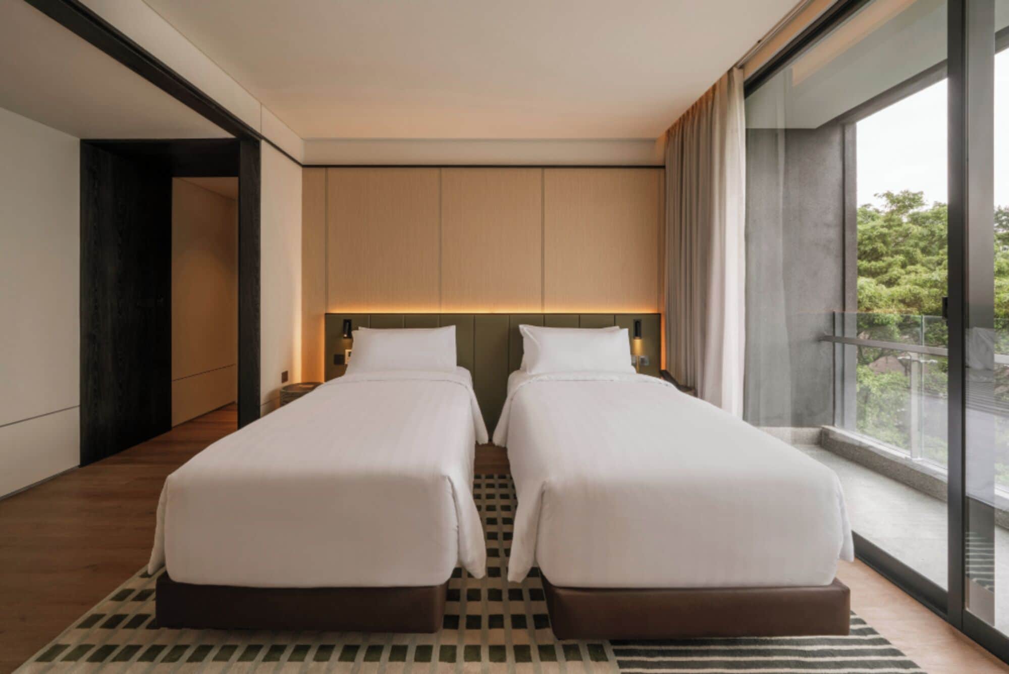 Une chambre d’hôtel moderne avec deux lits simples, une grande fenêtre donnant sur un paysage verdoyant et un tapis à motifs à carreaux.