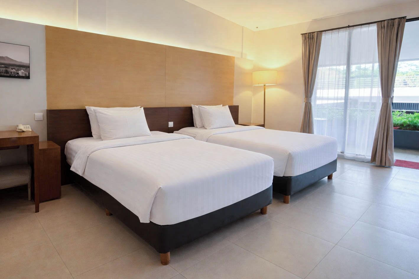A hotel room with two beds, a desk, a lamp, and a view of greenery through the window.