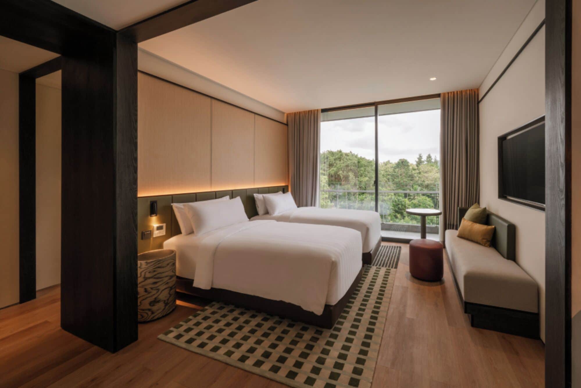 Habitación de hotel moderna con dos camas, un sofá, un televisor y un balcón con vistas a árboles.