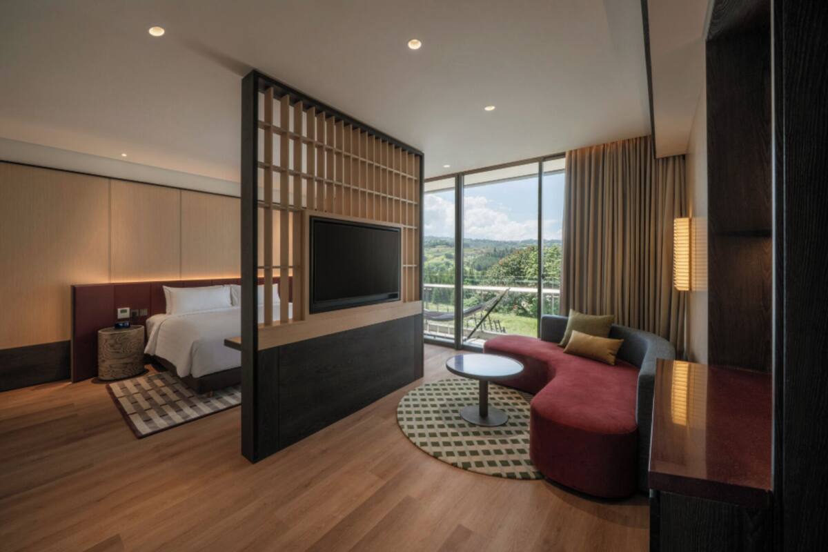 Una moderna camera d'albergo con un letto grande, una TV montata su un pannello di legno, un divano rosso e un balcone con vista sulla campagna.