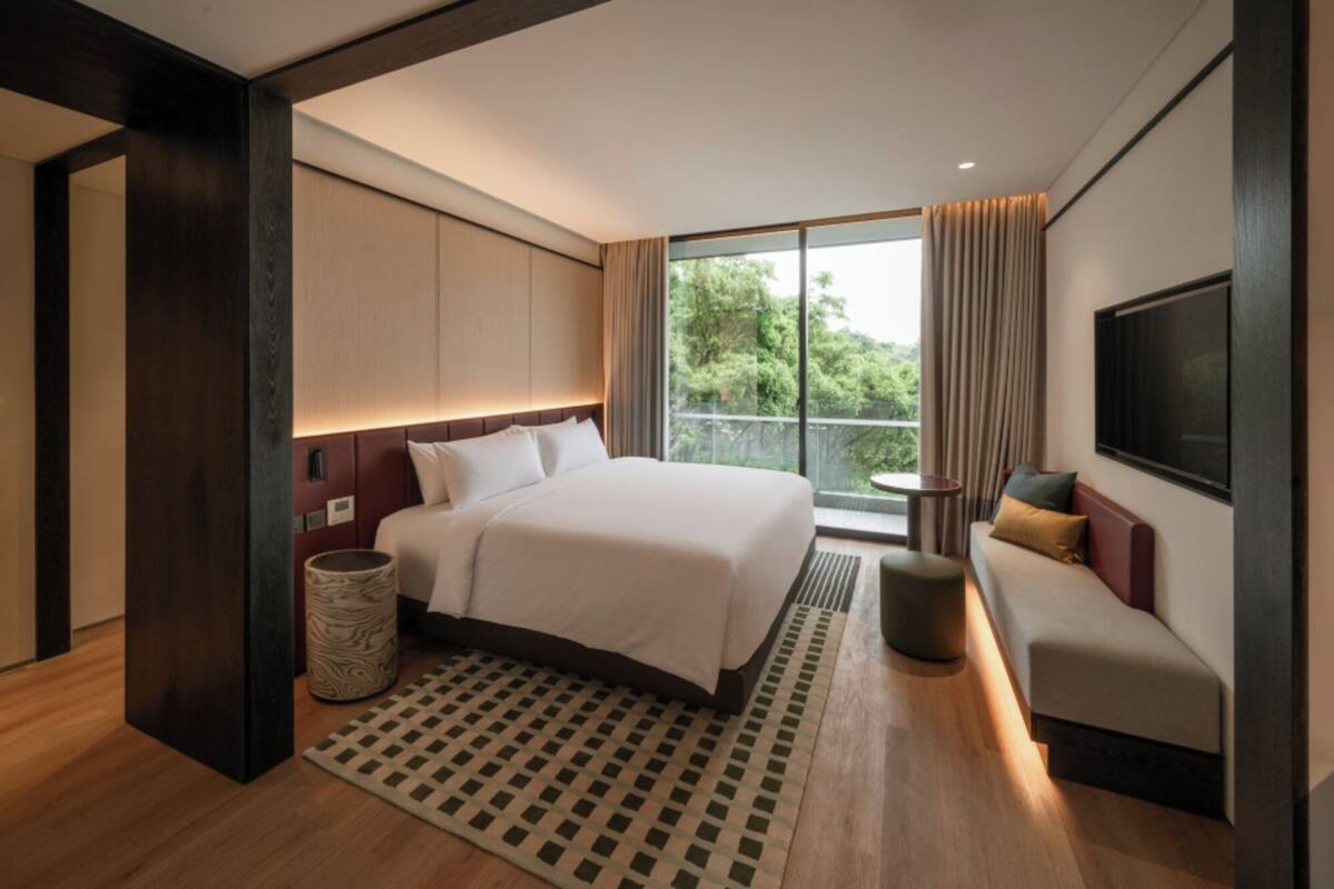 Una camera d'albergo moderna con un letto grande, una poltrona, una TV e un balcone con vista sugli alberi.