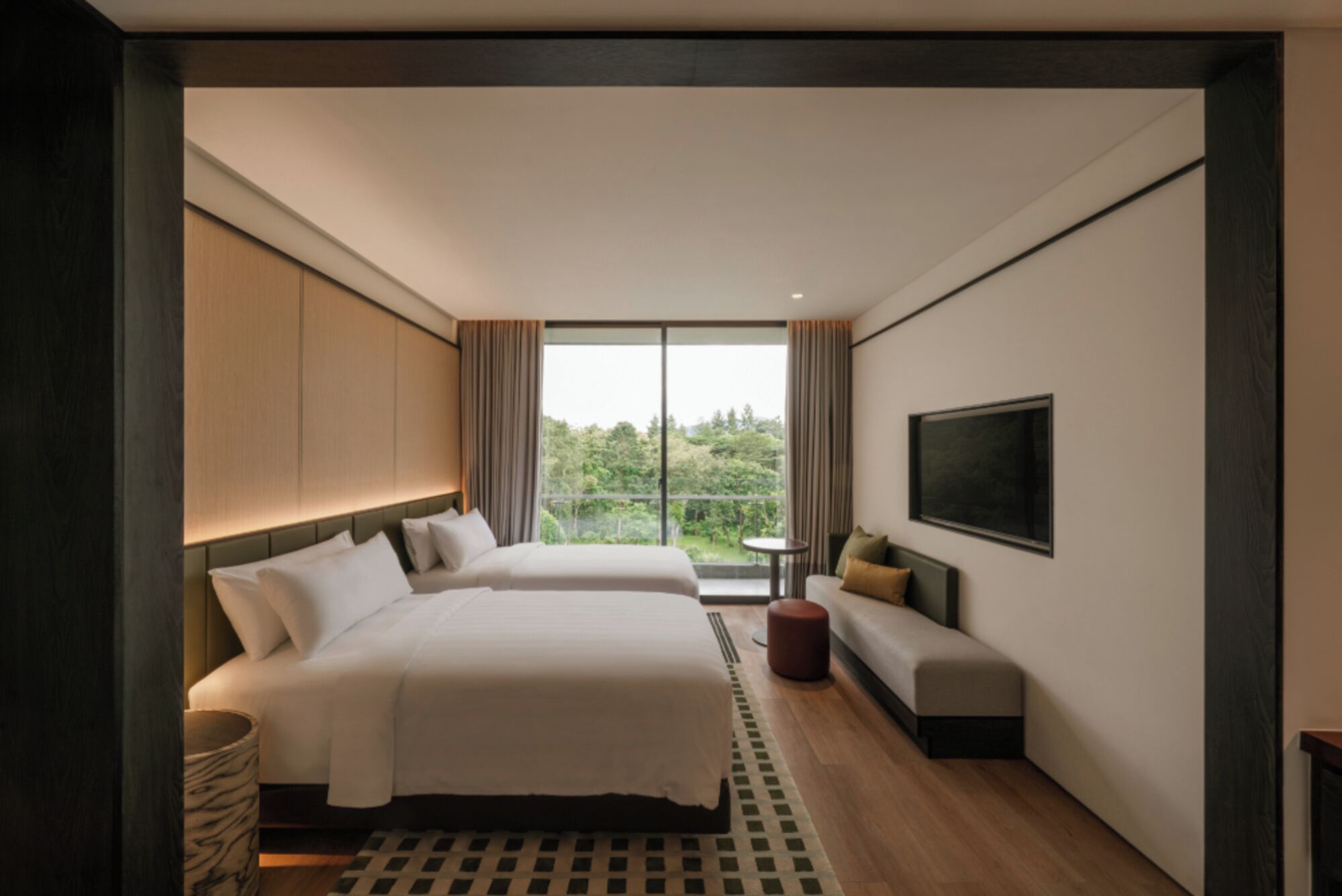 A hotel room with two beds, a sofa, a TV, and a view of greenery outside.