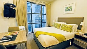 A hotel room with a bed, a desk, a chair, a TV, and a window with a view of buildings. - Quest Atrium Serviced Apartments (Wellington)