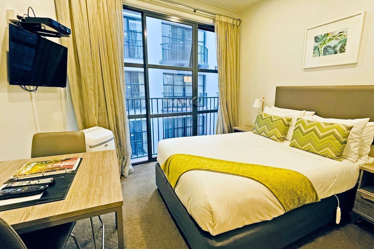 A hotel room with a bed, a desk, a chair, a TV, and a window with a view of buildings.