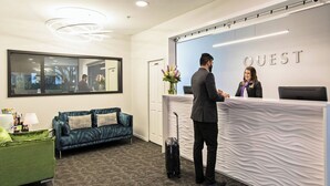 A hotel reception area with a reception desk, a blue sofa, a green armchair, and a large mirror on the wall. - Quest Atrium Serviced Apartments (Wellington)