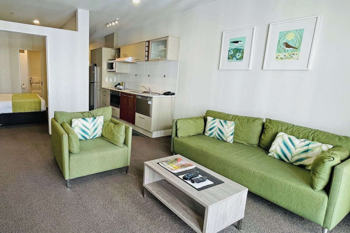 A modern living room with a green sofa, a matching armchair, and a coffee table. A kitchen is visible in the background with white cabinets and a microwave.