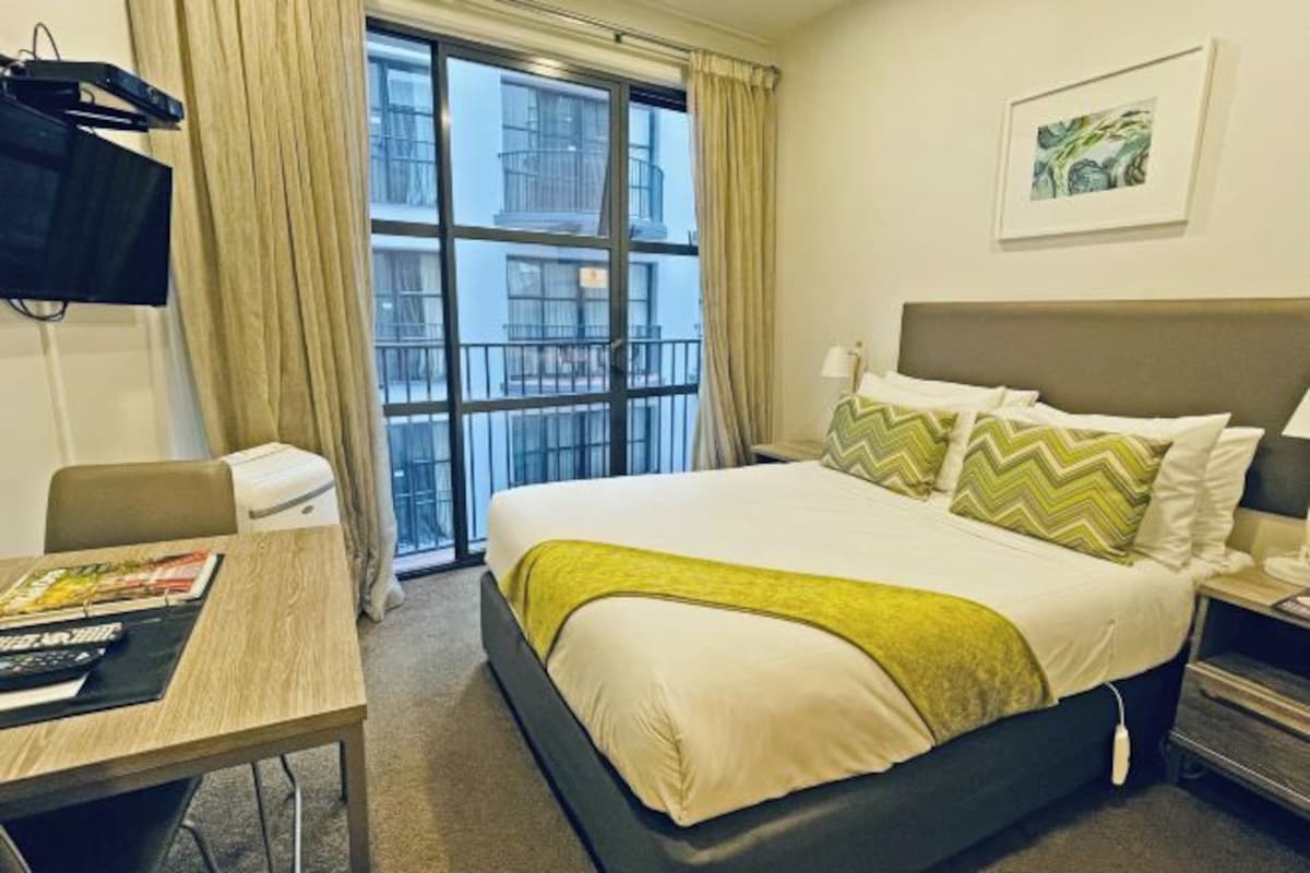 Una camera d'albergo con un letto, una scrivania, una sedia, una TV e una finestra con tende.