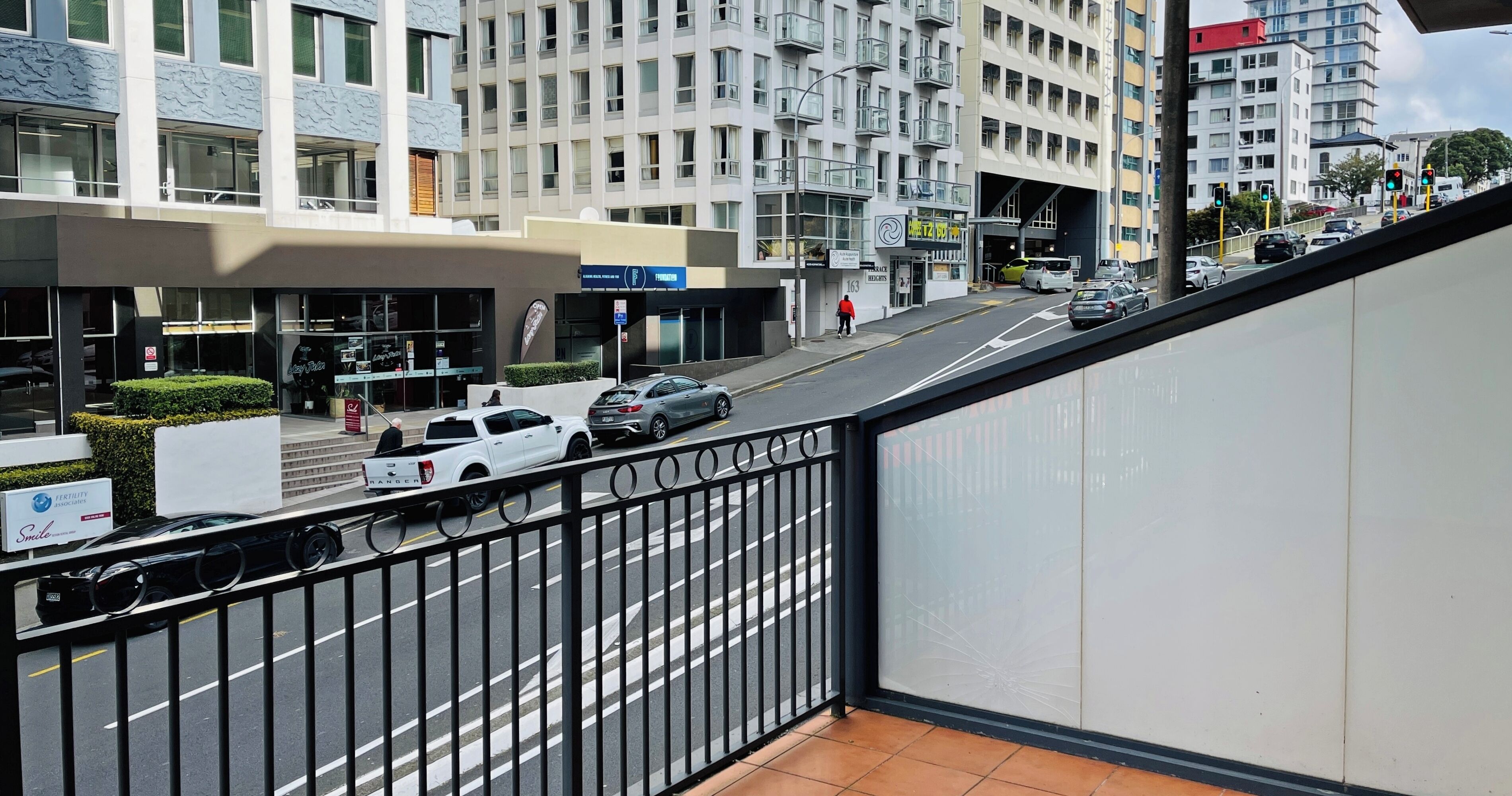 A modern building with multiple floors, a gated entrance, and a street with cars and a pedestrian.
