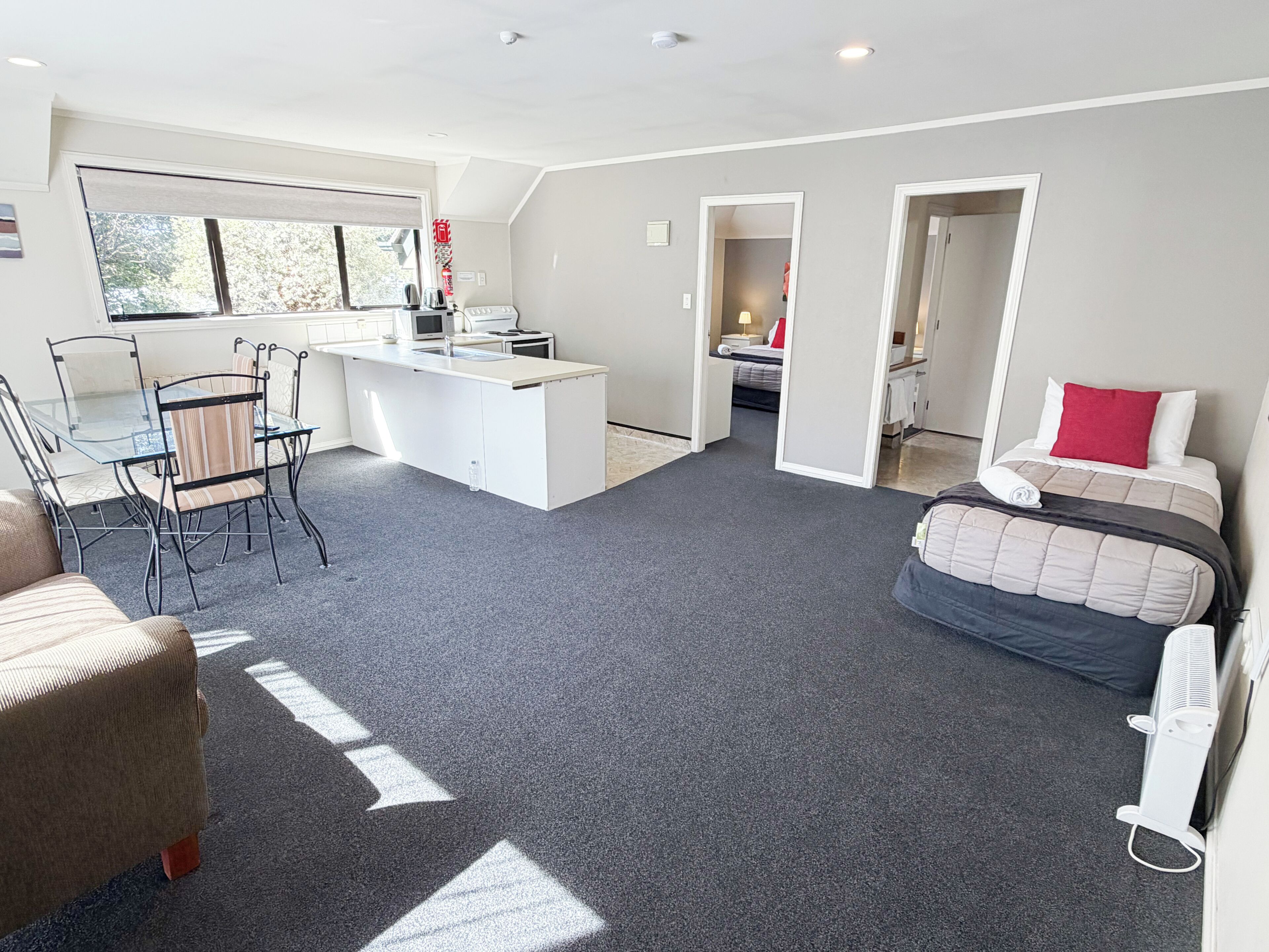 A modern hotel room with a kitchenette, a dining area, a bed with a red pillow, and a large window.