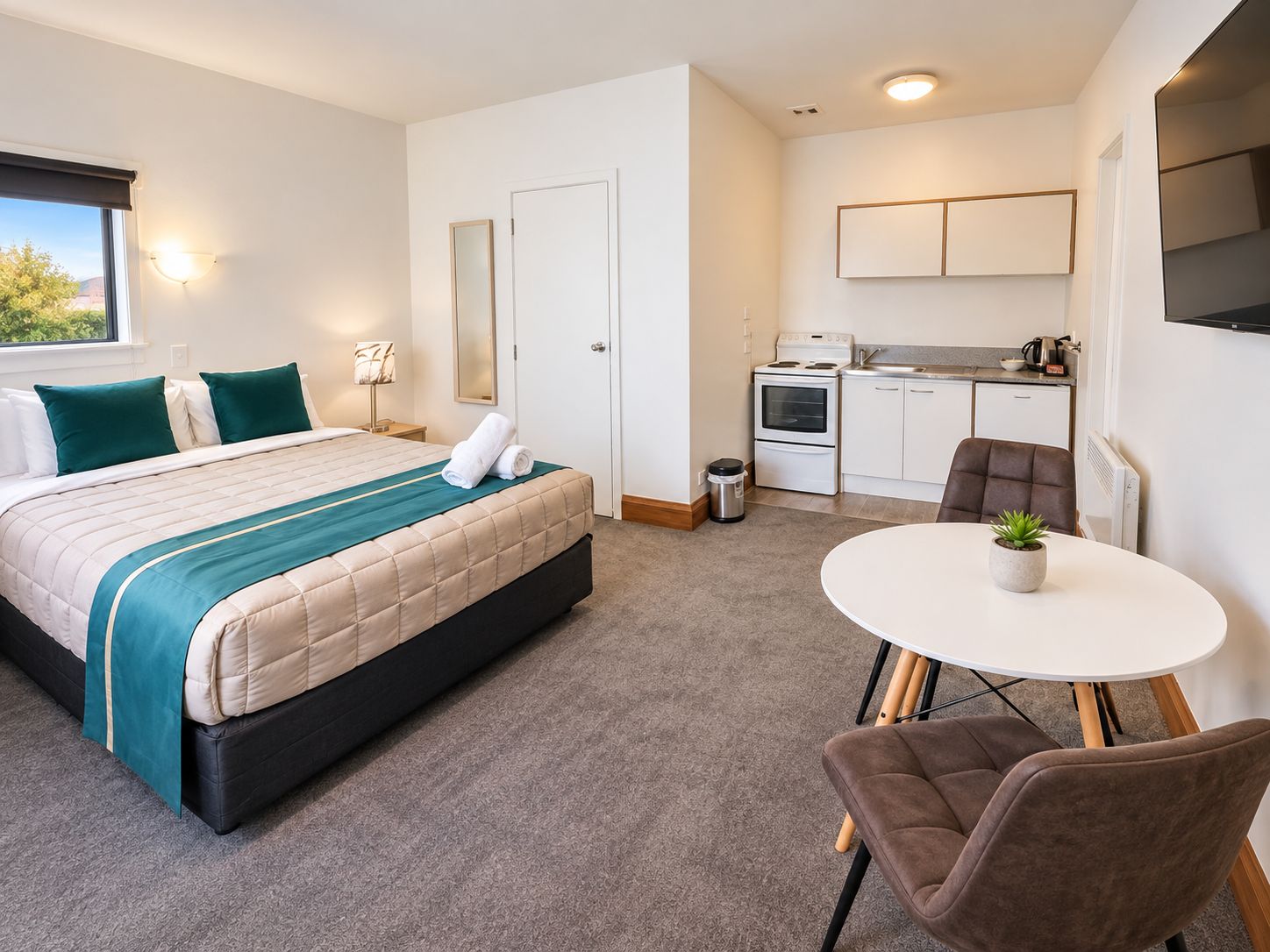 A compact hotel room with a bed, a small kitchenette, a dining table, and a chair.